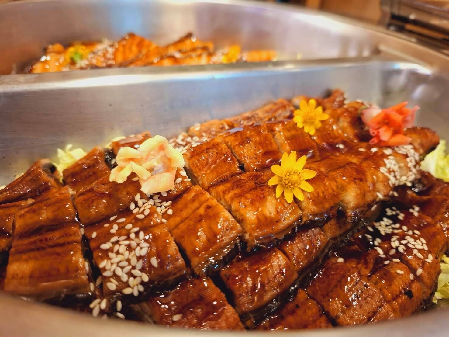 A large, glazed meat dish garnished with sesame seeds and flowers, likely served in a buffet setting.