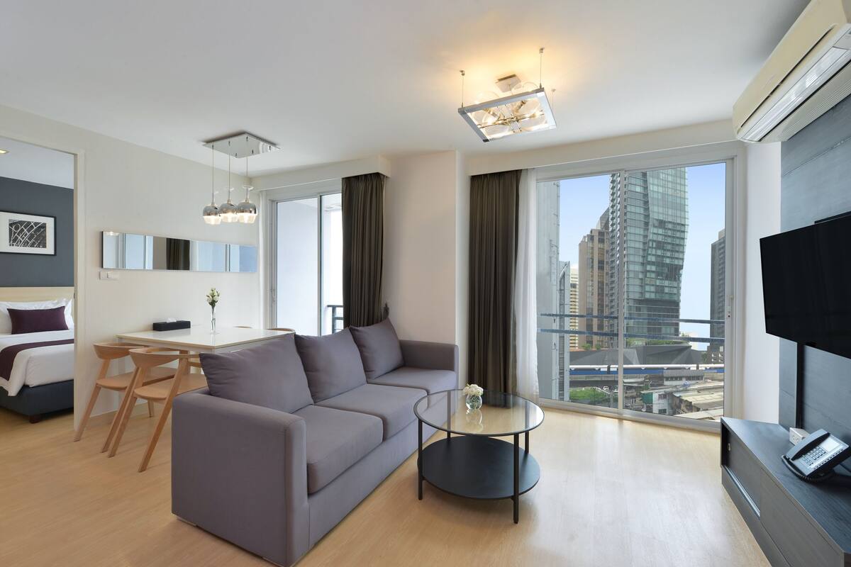 A modern living room with a grey sofa, a round coffee table, a dining area with a table and chairs, a flat-screen TV, and a large window with a city view.