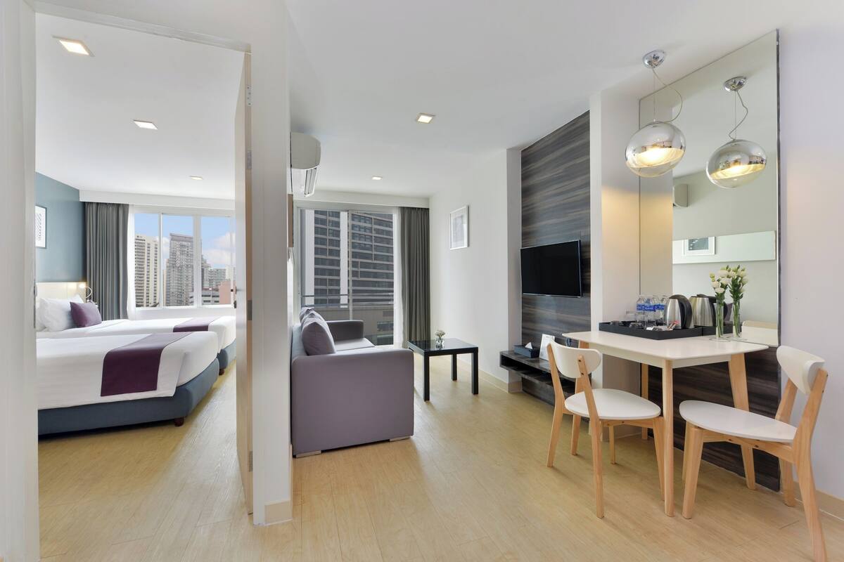 A modern hotel room with a bed, a sofa, a dining table with chairs, a TV, and a large window with a city view.