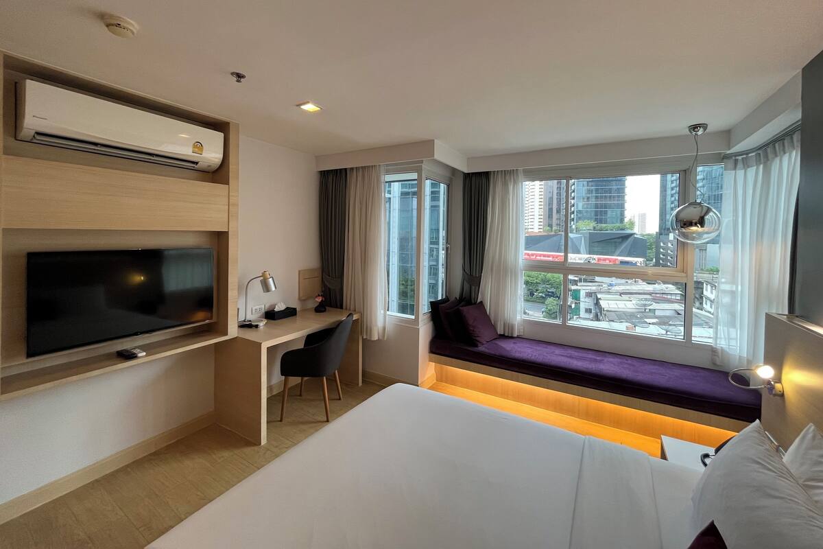 A hotel room with a bed, a desk, a TV, a window with a view of the city, and an air conditioning unit.