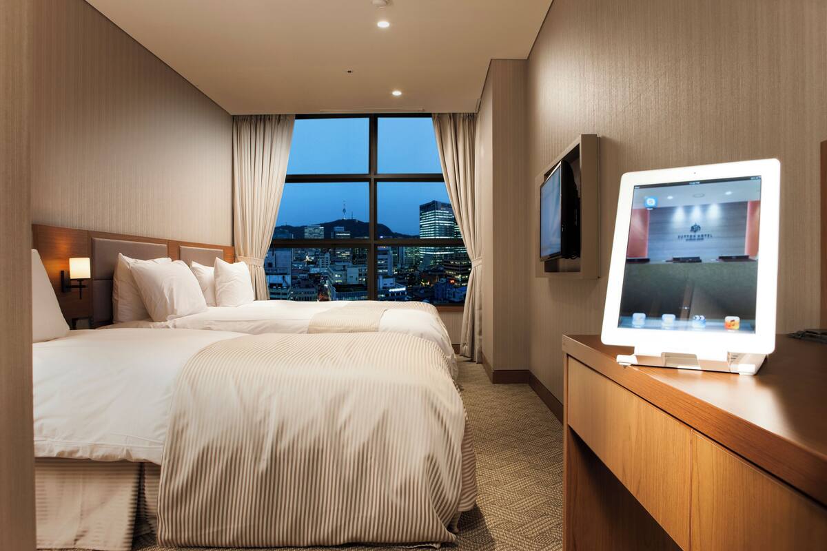 Habitación de hotel con una cama grande, un escritorio con una computadora y vistas a un paisaje urbano a través de la ventana.