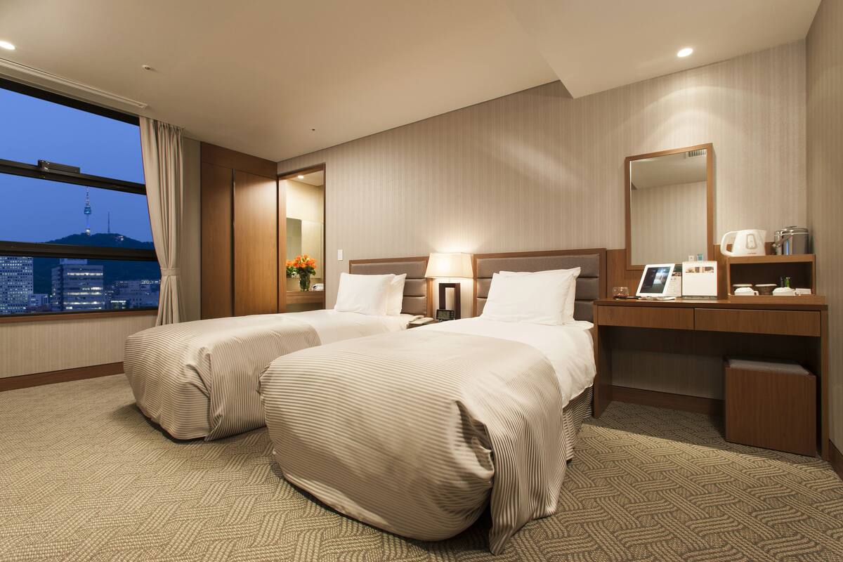 Habitación de hotel con dos camas, un escritorio, un espejo y vista a un paisaje urbano al anochecer.