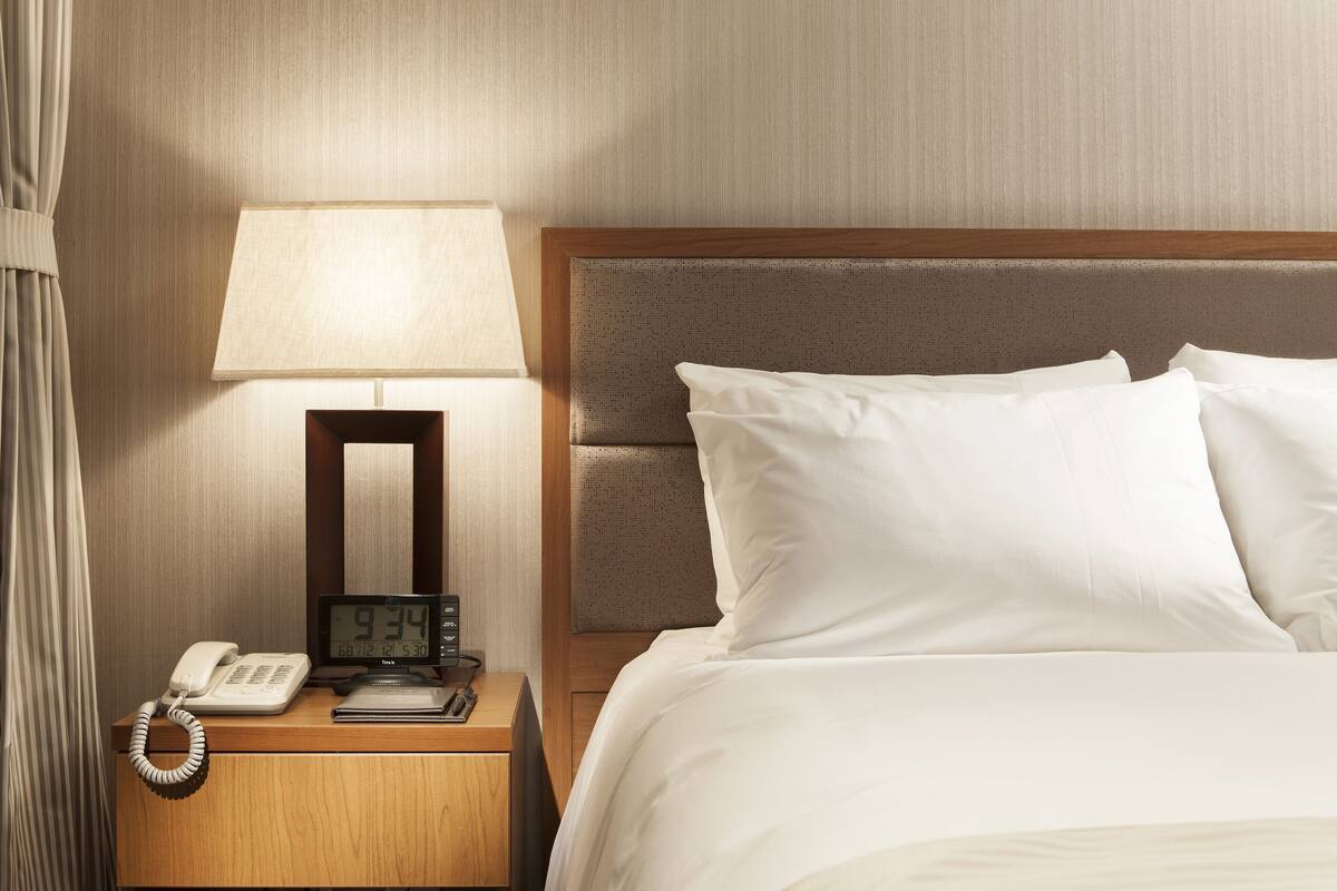 Habitación de hotel con mesita de noche de madera, lámpara, reloj digital, teléfono y cama bien tendida con sábanas blancas.