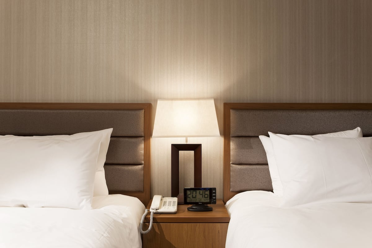 Habitación de hotel con dos camas, una mesita de noche con lámpara, un reloj digital y un teléfono.