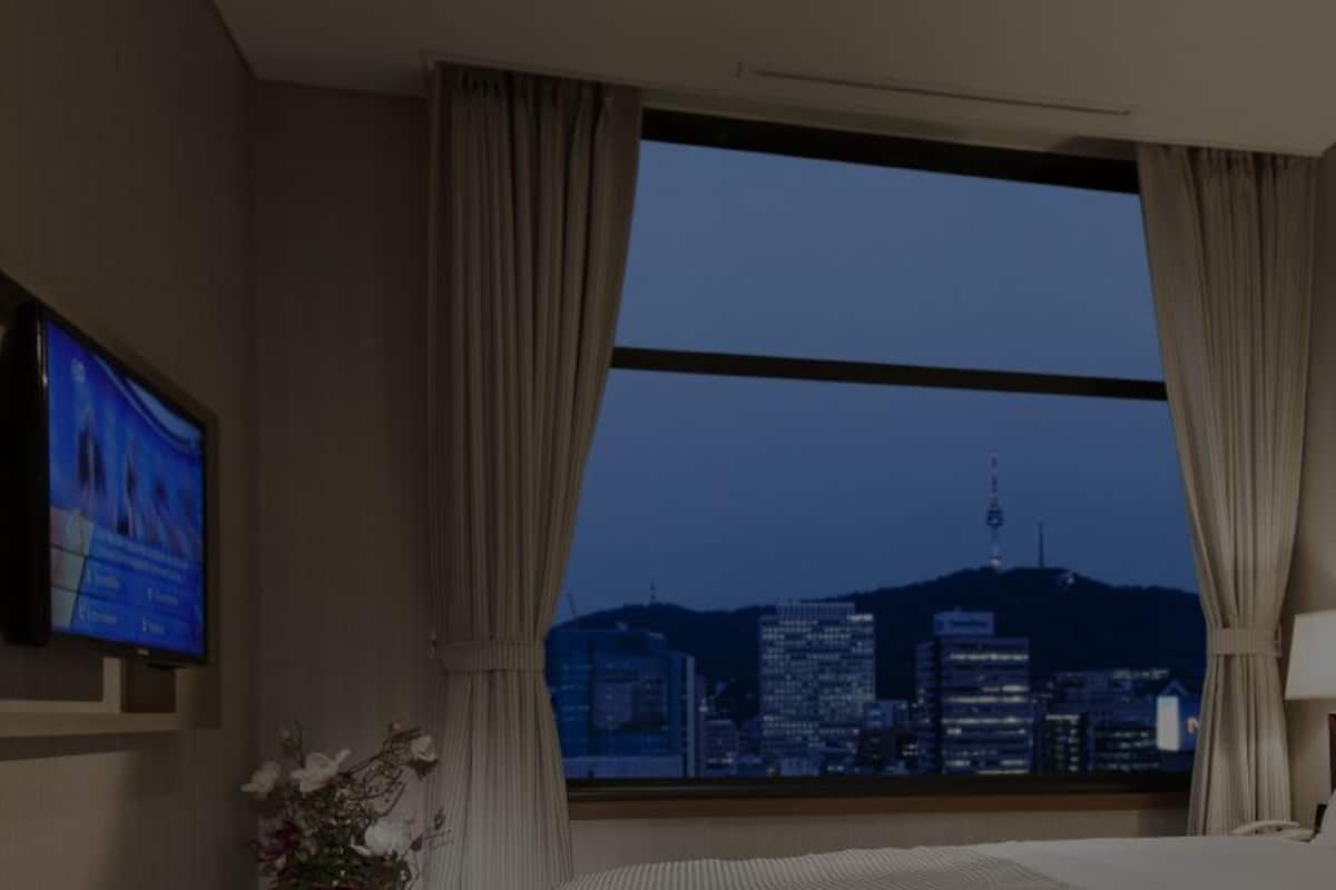 Habitación de hotel con televisor montado en la pared, un ventanal con cortinas y vistas a un paisaje urbano nocturno.