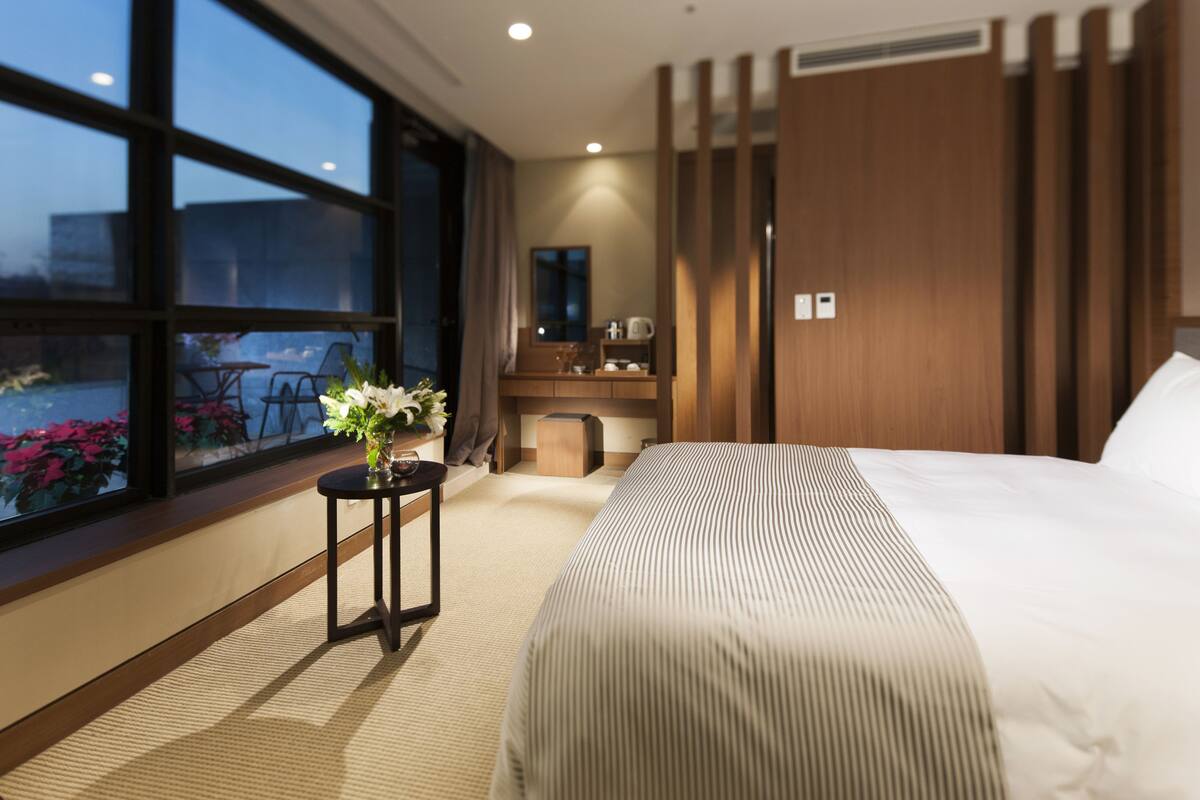 Habitación de hotel con un ventanal grande, una cama con ropa de cama a rayas, una mesita con un florero, un escritorio con espejo y un panel de pared de madera.