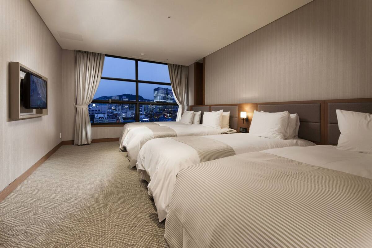 Habitación de hotel con tres camas, un ventanal grande con vista a la ciudad, un televisor de pantalla plana montado en la pared y piso alfombrado.