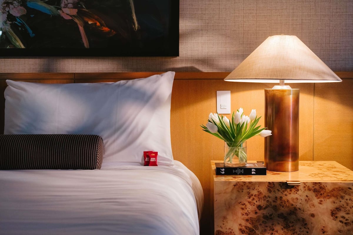 A neatly made bed with white linens, a striped pillow, a red item on the nightstand, a vase with white flowers, and a lamp on a wooden side table.