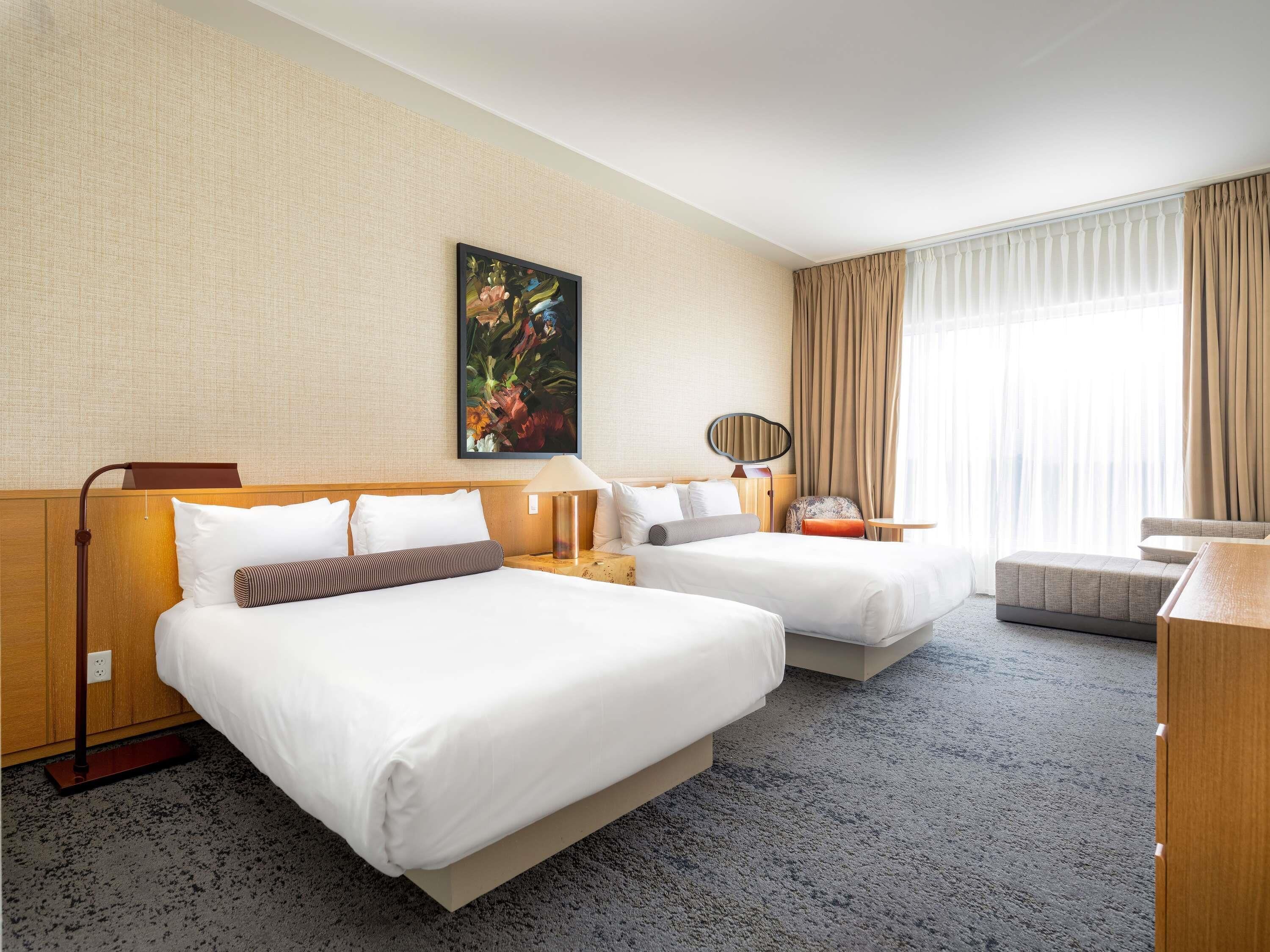 A hotel room with two beds, a desk, a chair, a painting on the wall, and a window with curtains.