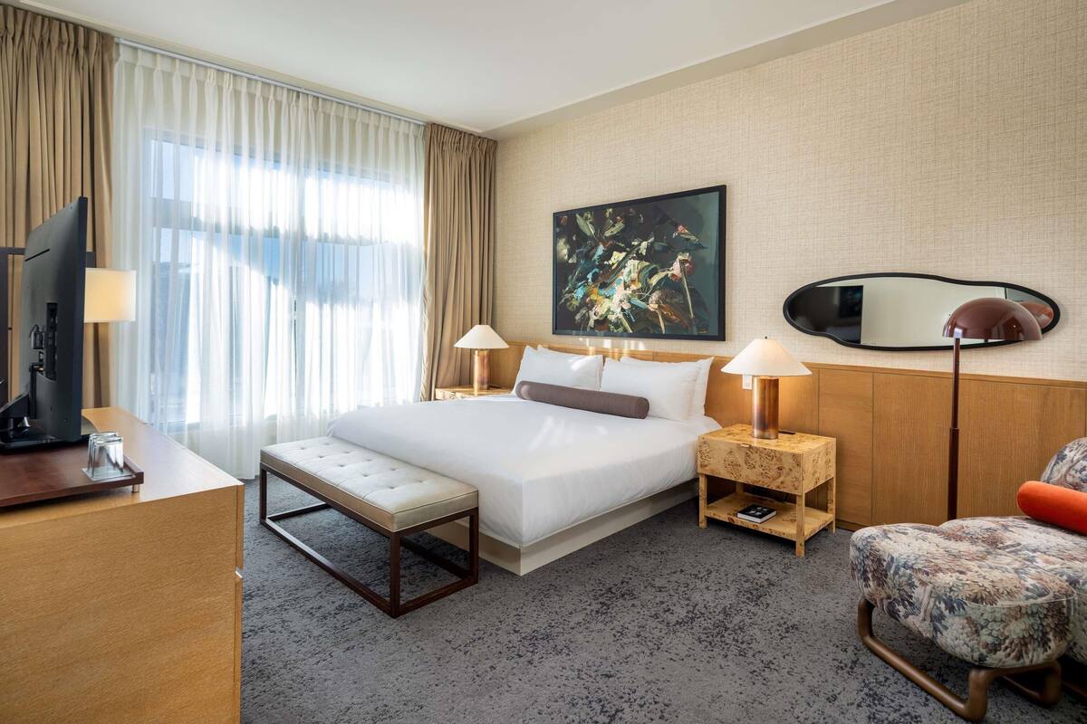 A hotel room with a bed, bedside tables, a desk, a television, a chair, and a large painting on the wall.