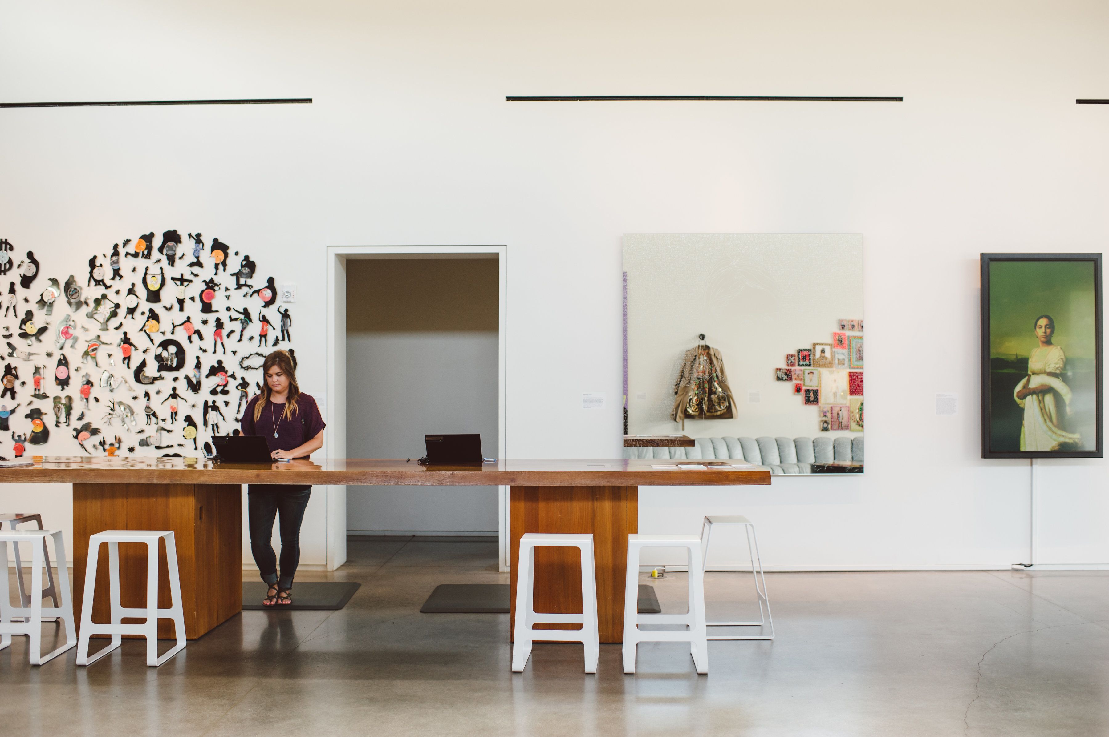 A modern art gallery with a central reception desk, white stools, and a variety of artworks on display.