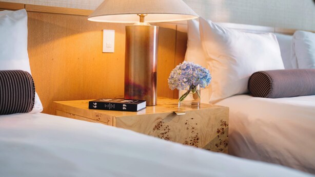 Un lit bien fait, avec du linge de lit blanc, une table de chevet avec une lampe, un livre et un vase rempli de fleurs.