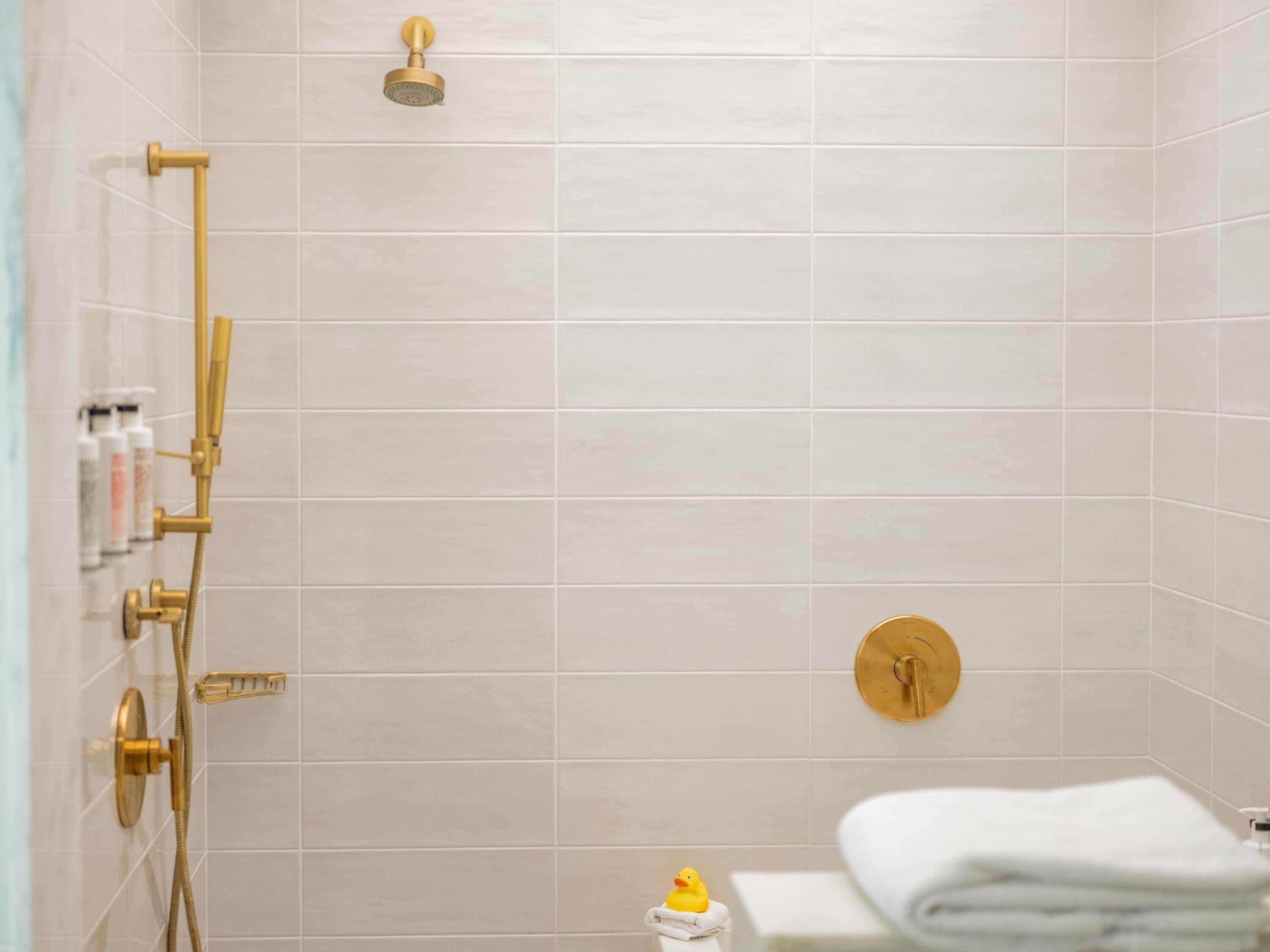 A shower with gold fixtures, a white tiled wall, and a small rubber duck.