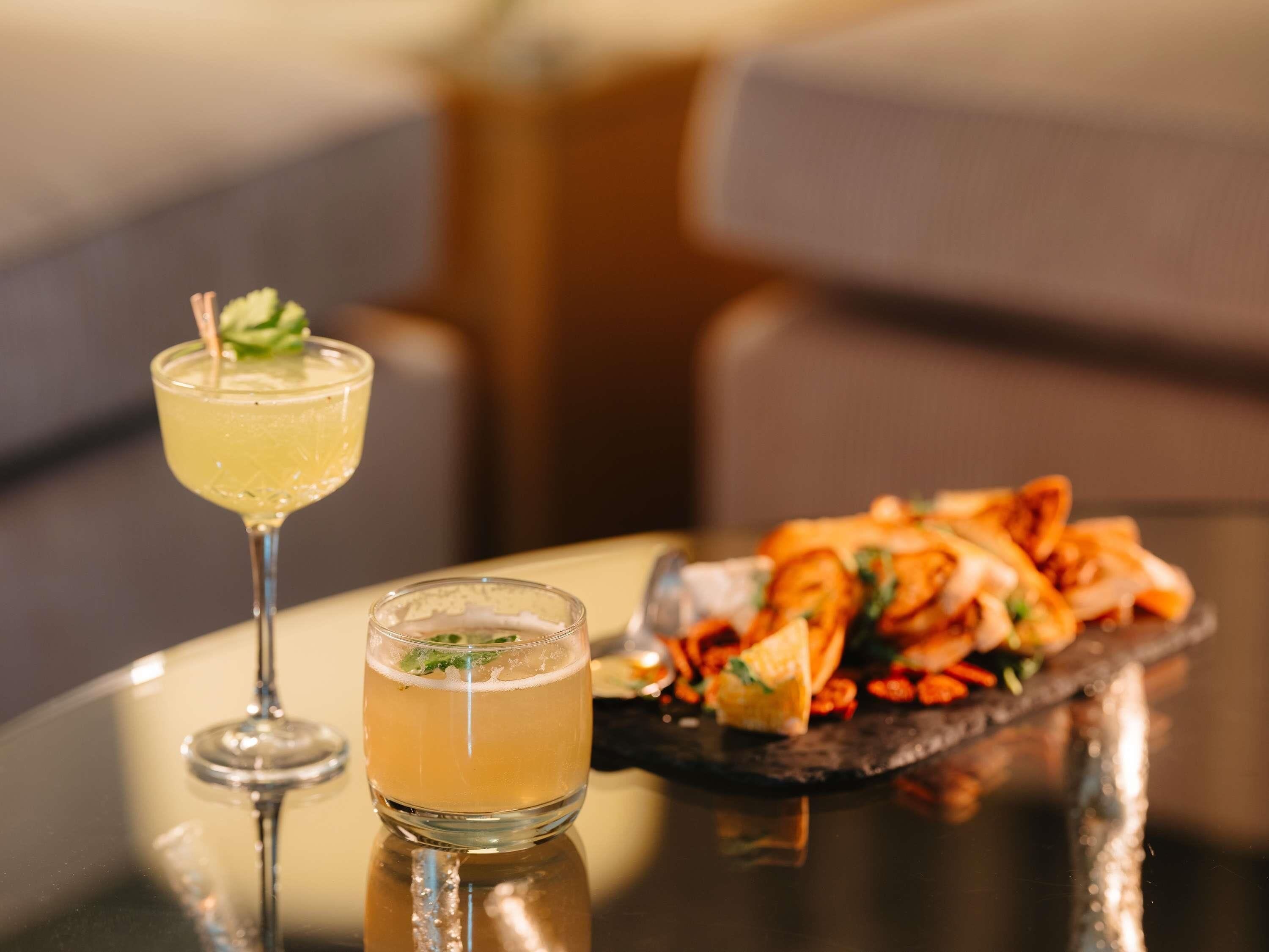 A cocktail glass and a small glass of water on a reflective surface with a plate of food in the background.