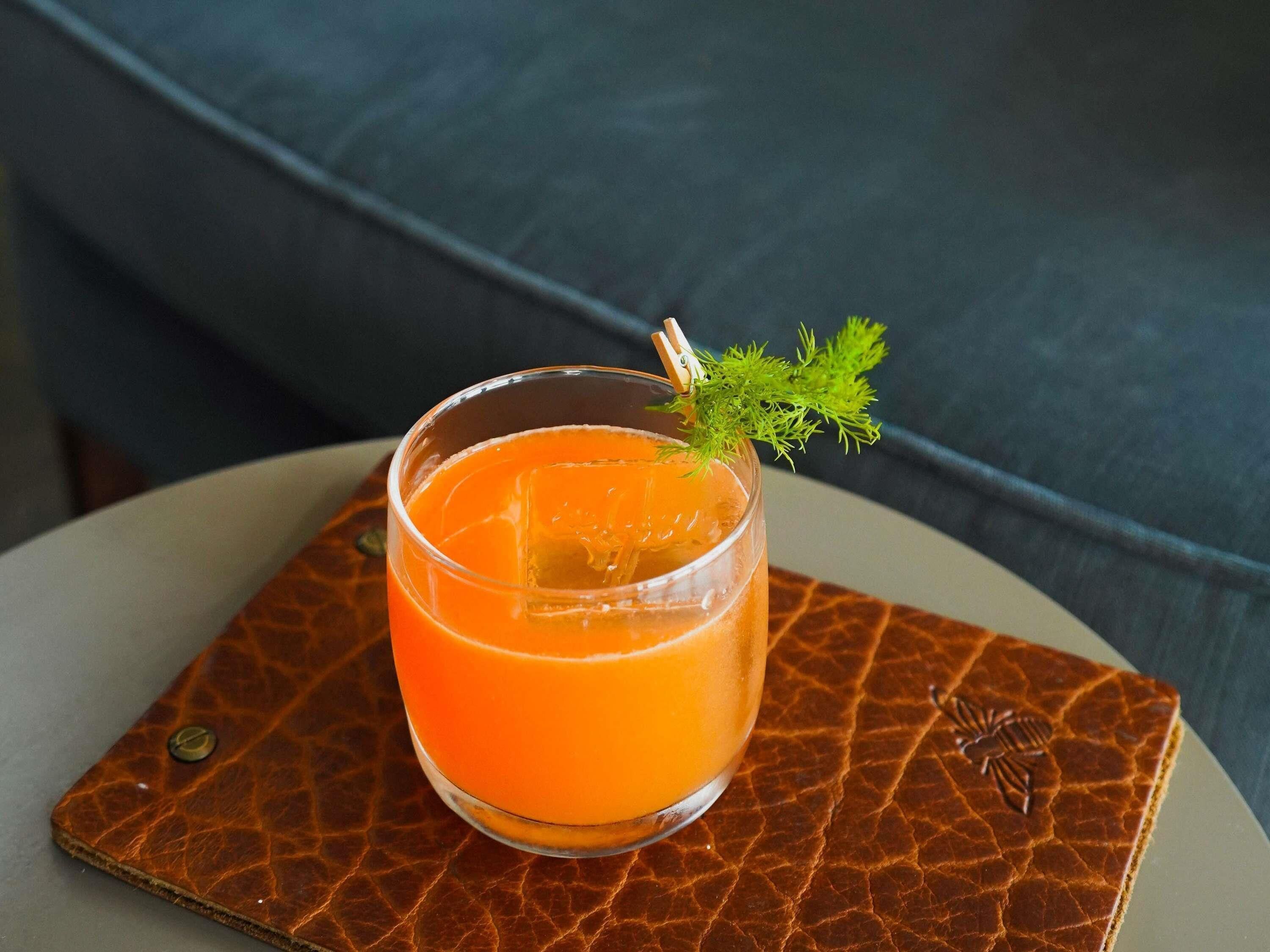 A glass of orange juice with a garnish on a textured coaster, placed on a round table.