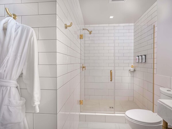 Un baño moderno con azulejos blancos, ducha con mampara de vidrio, inodoro y una bata colgada en un perchero.