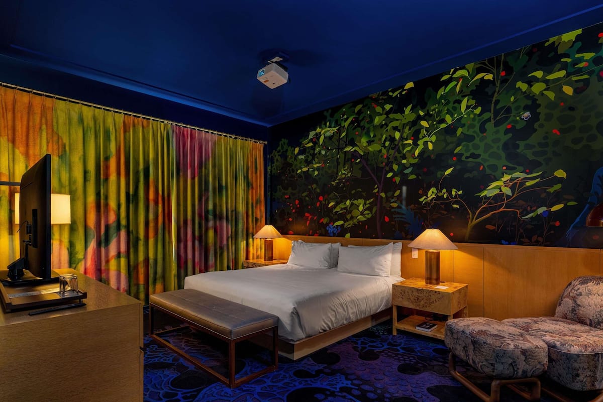 A hotel room with a bed, two bedside tables with lamps, a desk with a computer, a bench, a chair, and a large mural of a forest.