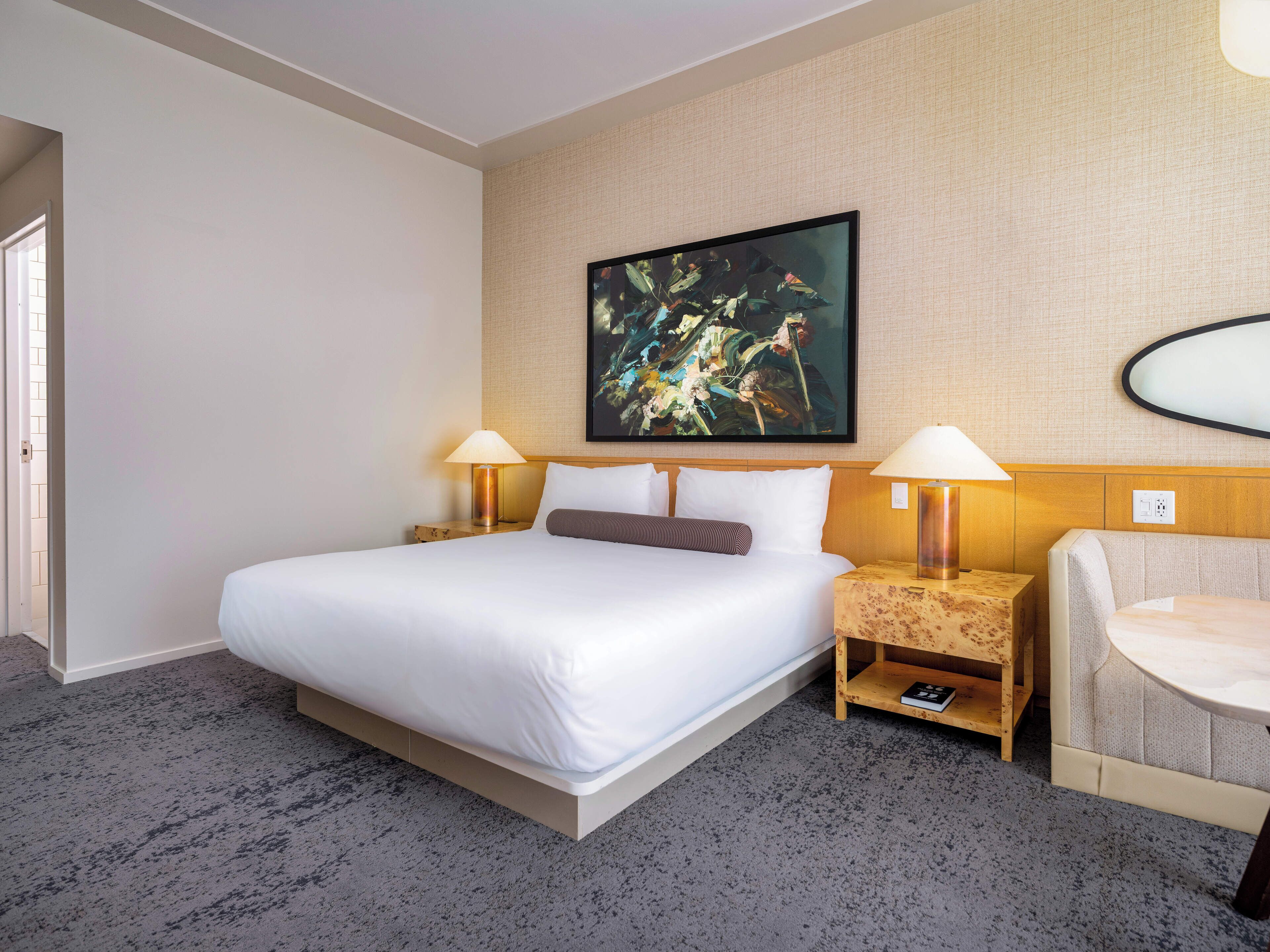A hotel room with a large bed, two bedside tables with lamps, a wooden nightstand, a chair, and a large abstract painting on the wall.