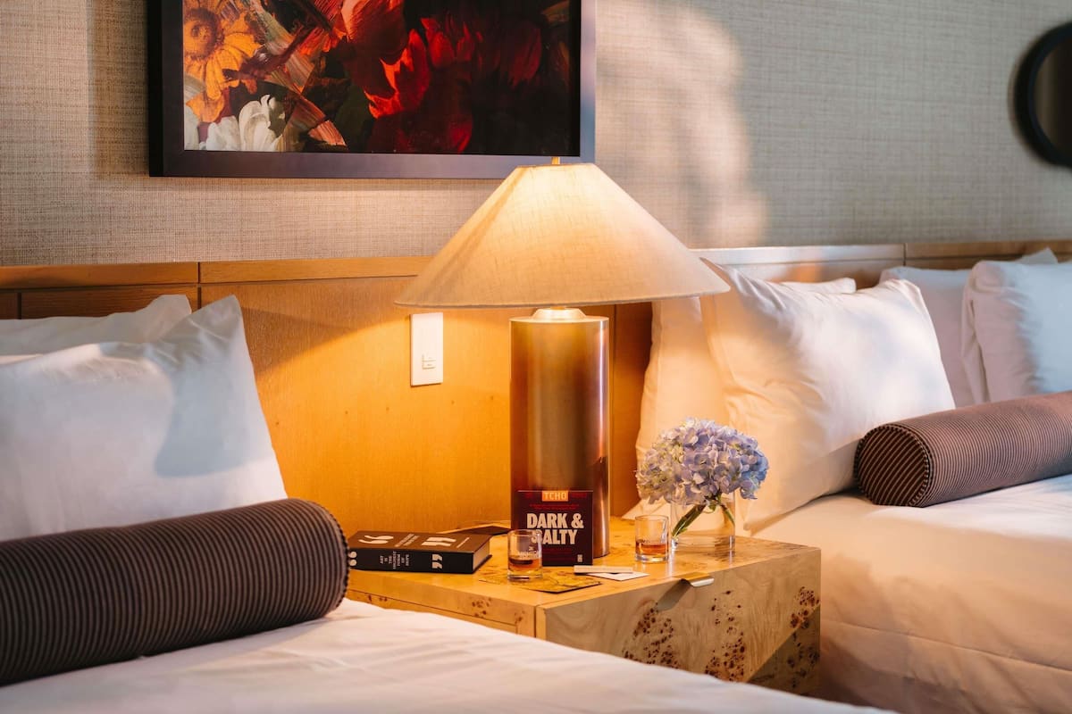A hotel room with a bed, a bedside table with a lamp, a book, and a bottle of Dark & Salty.