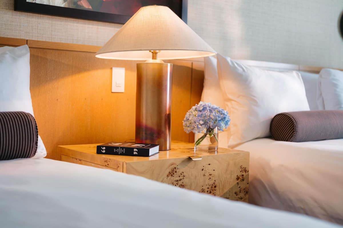A neatly made hotel bed with a lamp, a book, and a vase with flowers on a wooden side table.