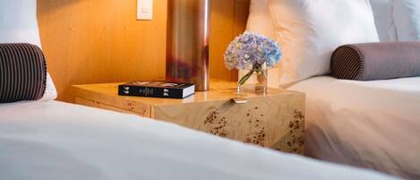 Una cama de hotel bien hecha, con una lámpara, un libro y un jarrón con flores sobre una mesita de madera.