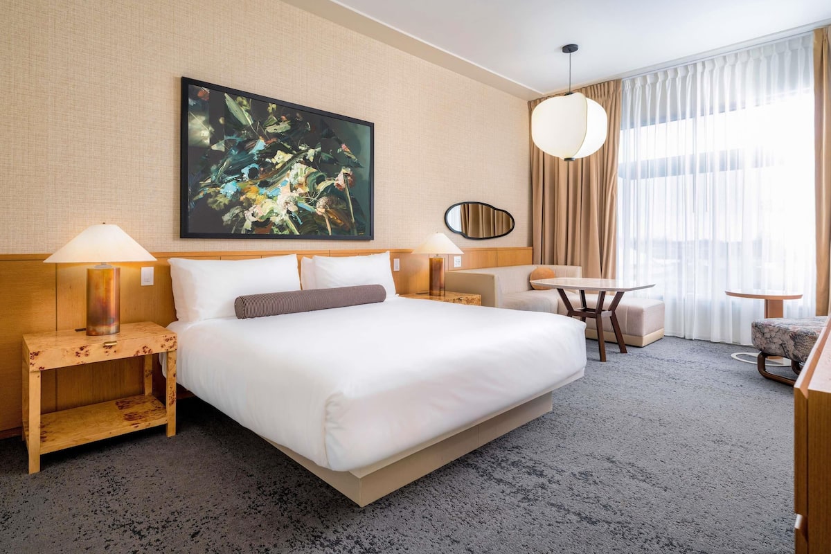 A hotel room with a large bed, a desk, a chair, a lamp, a painting, and a window with curtains.