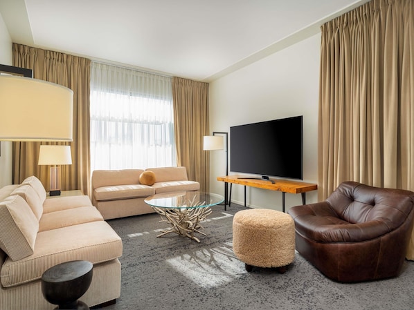 A hotel room with a sofa, ottoman, coffee table, TV, and curtains.