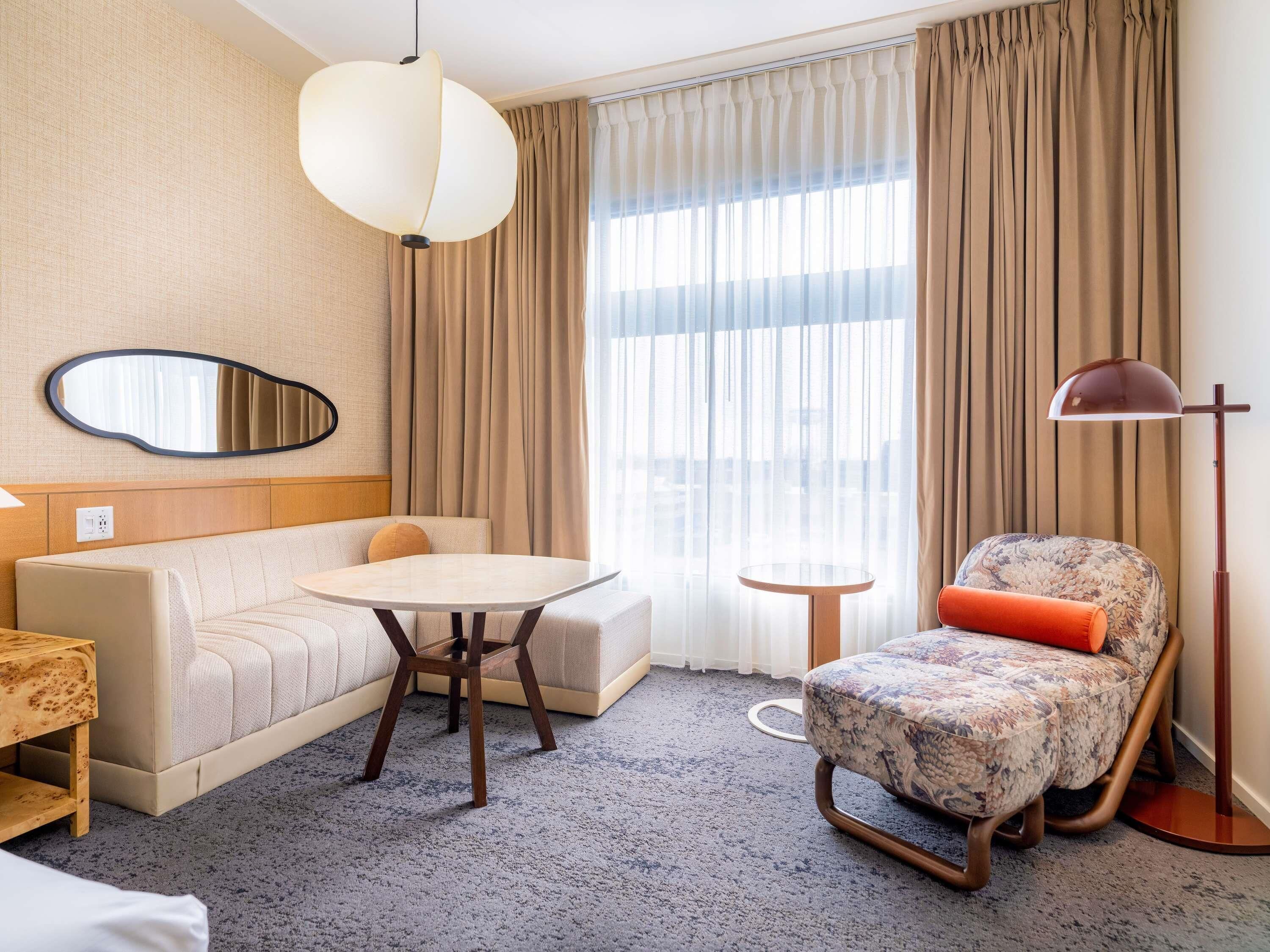 Ett hotellrum med en soffa, ett runt bord, en mönstrad fåtölj, en golvlampa, en spegel och ett stort fönster med gardiner.