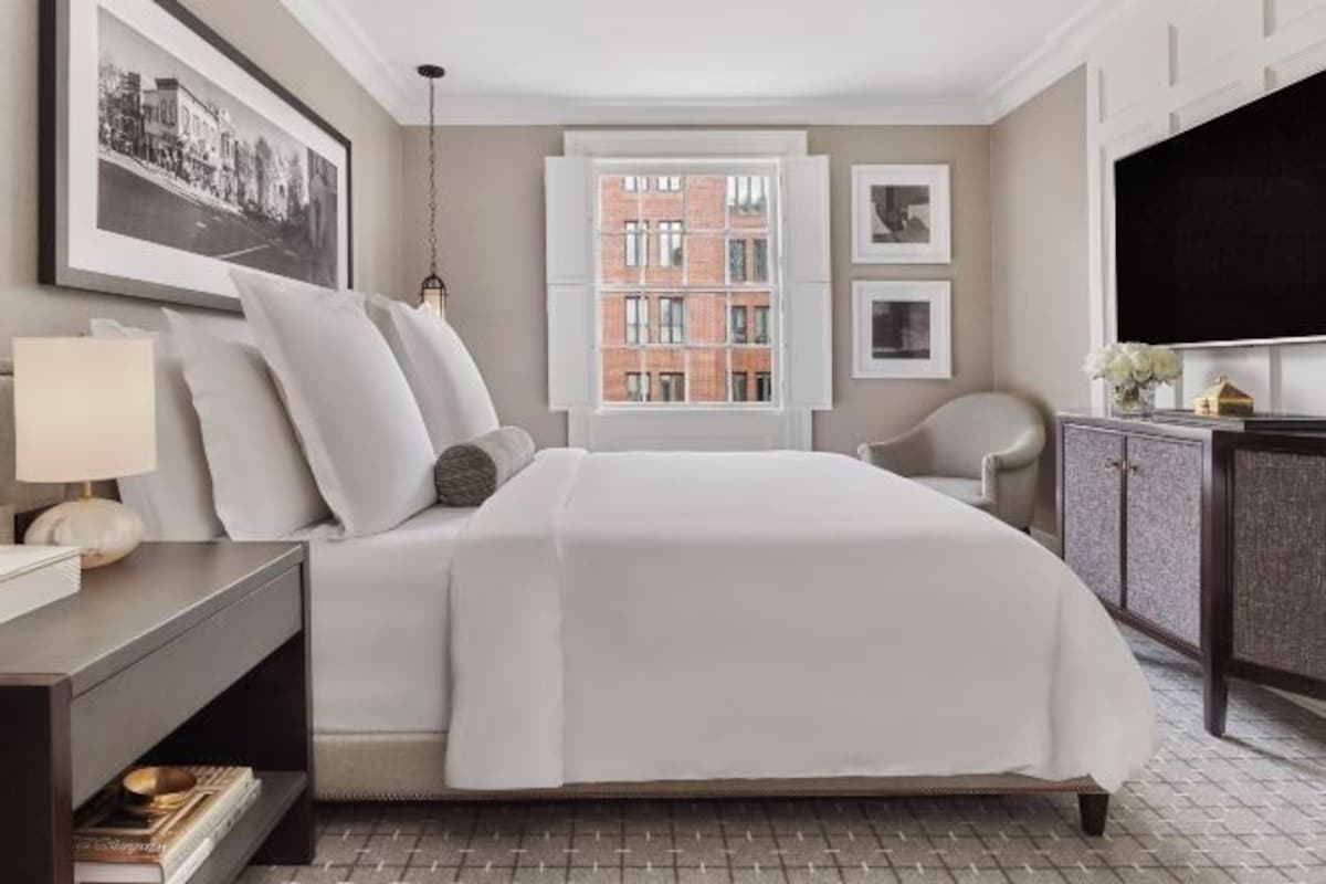 A hotel room with a large bed, a desk with a lamp, a chair, a TV, and a window with a cityscape view.