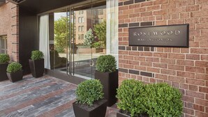 Ein moderner Gebäudeeingang mit Ziegelverkleidung, einer Glastür und einem Schild mit der Aufschrift 'Rosewood Washington, DC'. Übertöpfe und ein gemusterter Boden sind ebenfalls vorhanden.