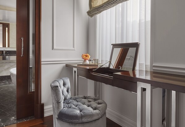 A modern bathroom with a marble floor, a wooden door, a grey tufted chair, and a vanity with a mirror and sink.