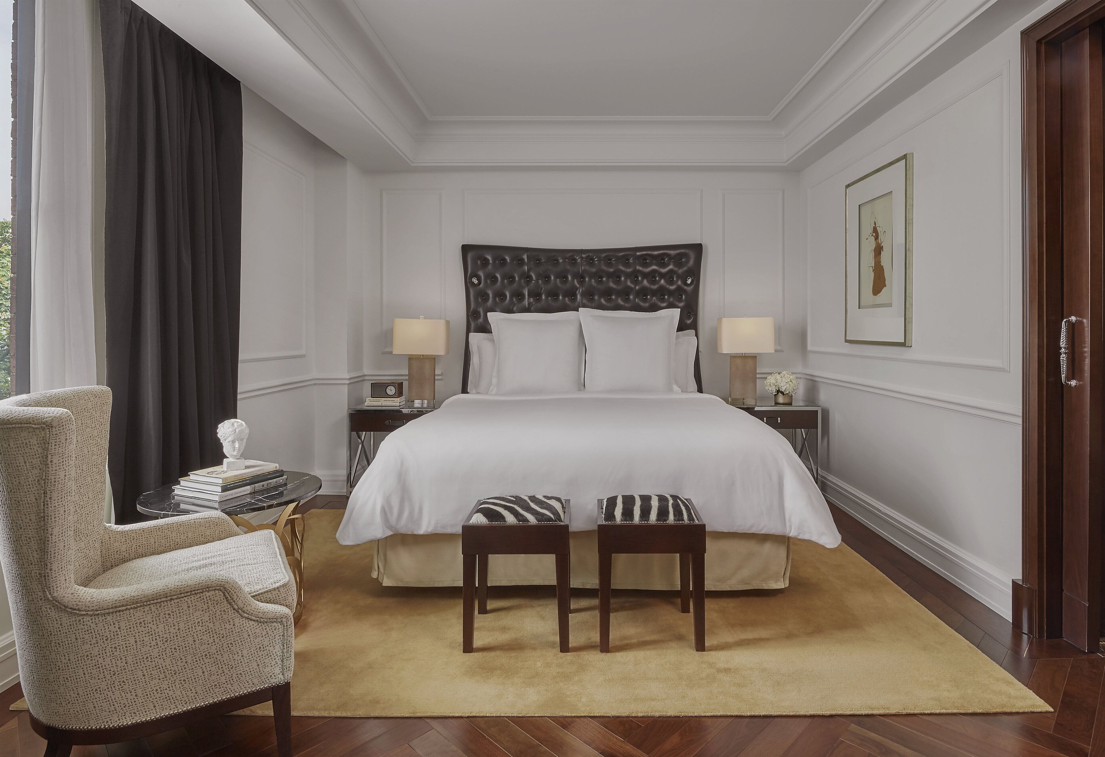 A neatly arranged bedroom with a large bed, two bedside tables with lamps, a chair, and a small table with books and a bust.