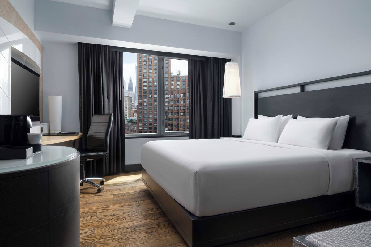 Habitación de hotel con una cama grande, un escritorio con una silla, una cafetera y vistas a edificios altos.