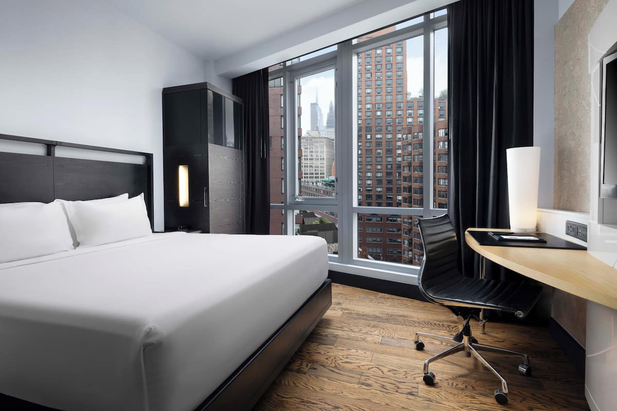 Una habitación de hotel moderna con una cama grande, un escritorio con lámpara y vistas a edificios de la ciudad a través de amplias ventanas.