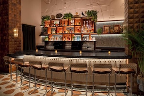 Una zona de bar con mostrador de mármol, taburetes, estantes con botellas y plantas decorativas.