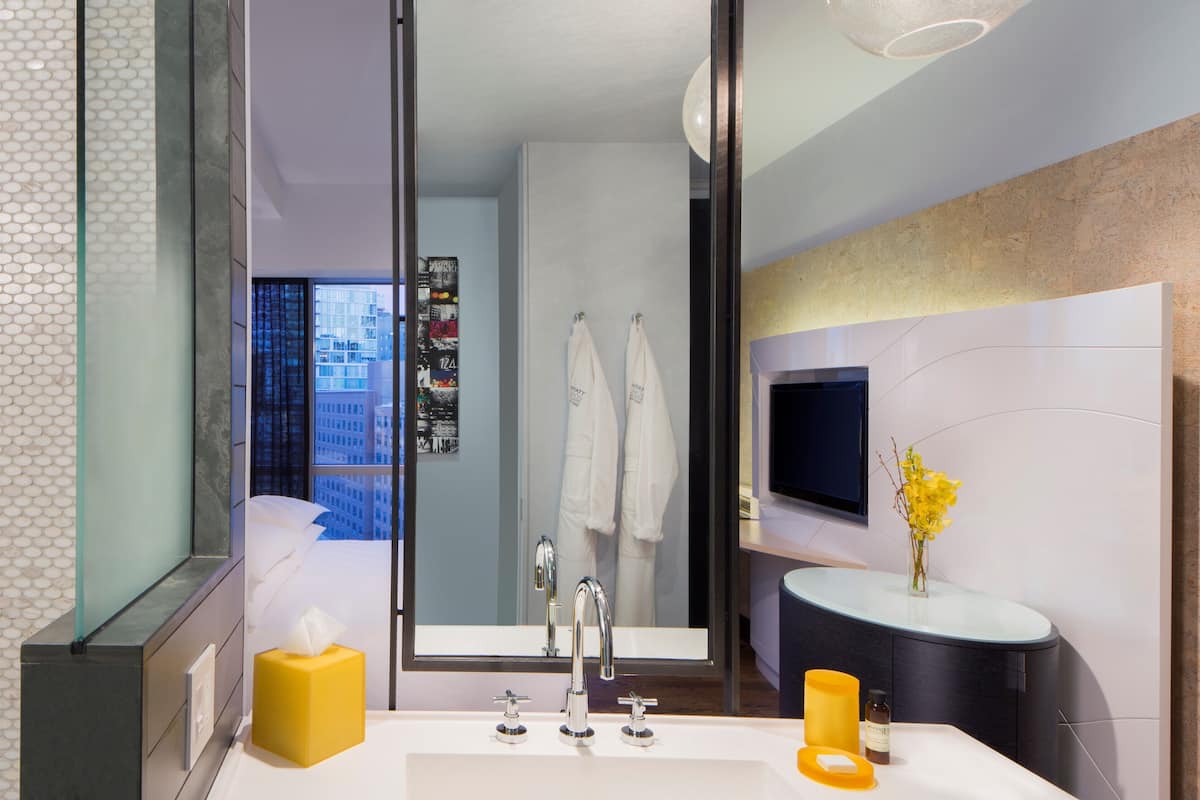 Un baño moderno con un espejo grande, un lavamanos y un inodoro. Hay una ventana con vistas a la ciudad, y toallas colgadas en la pared.