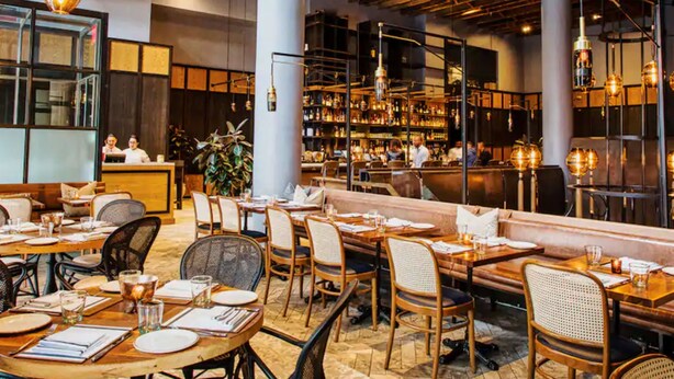 A restaurant interior with wooden tables, cushioned seating, hanging pendant lights, and a bar area with shelves of bottles.