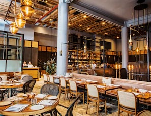 Interior de un restaurante con mesas de madera, asientos acolchados, lámparas colgantes y una zona de bar con estanterías llenas de botellas.