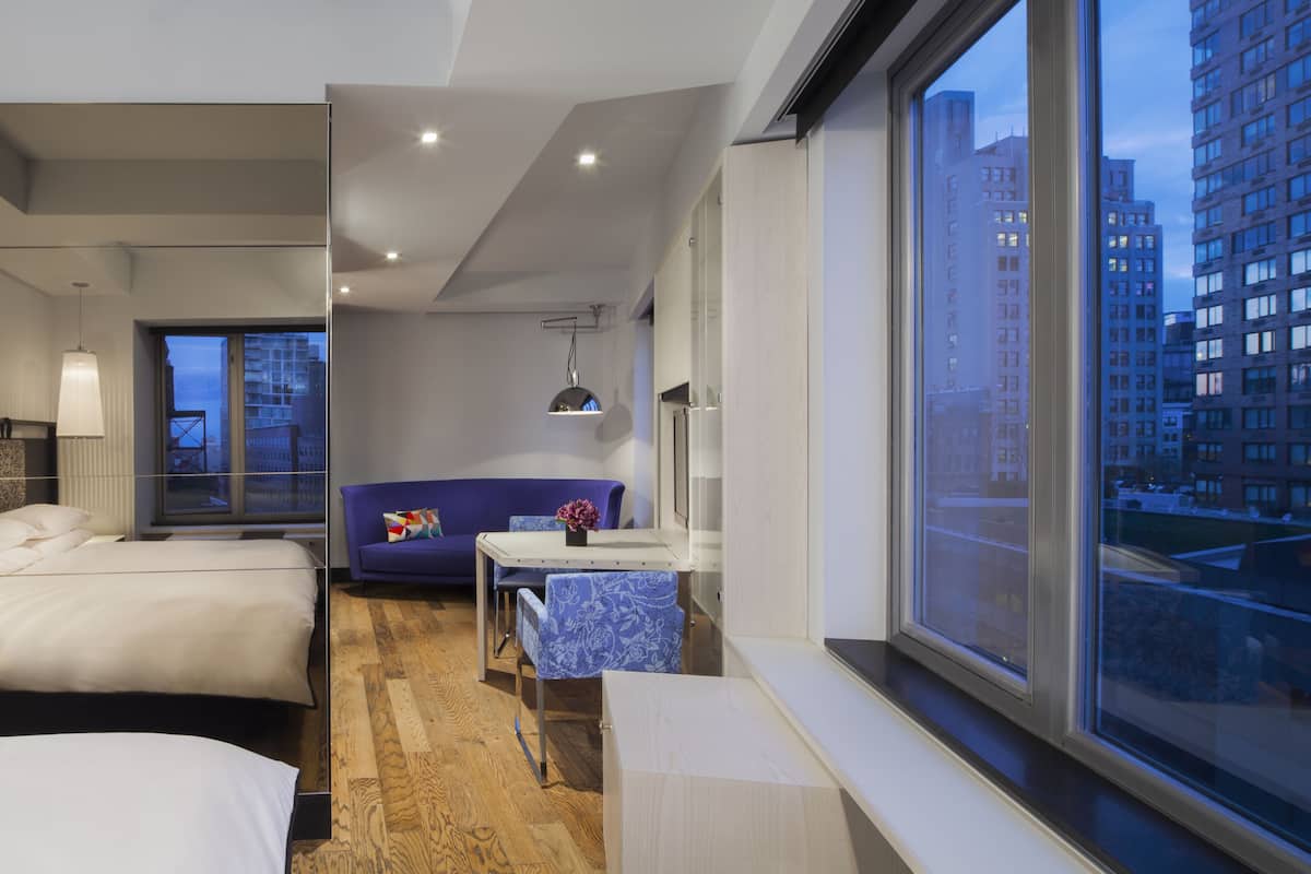 Una habitación de hotel moderna con un ventanal grande, una cama, un sofá, una mesita y un escritorio.