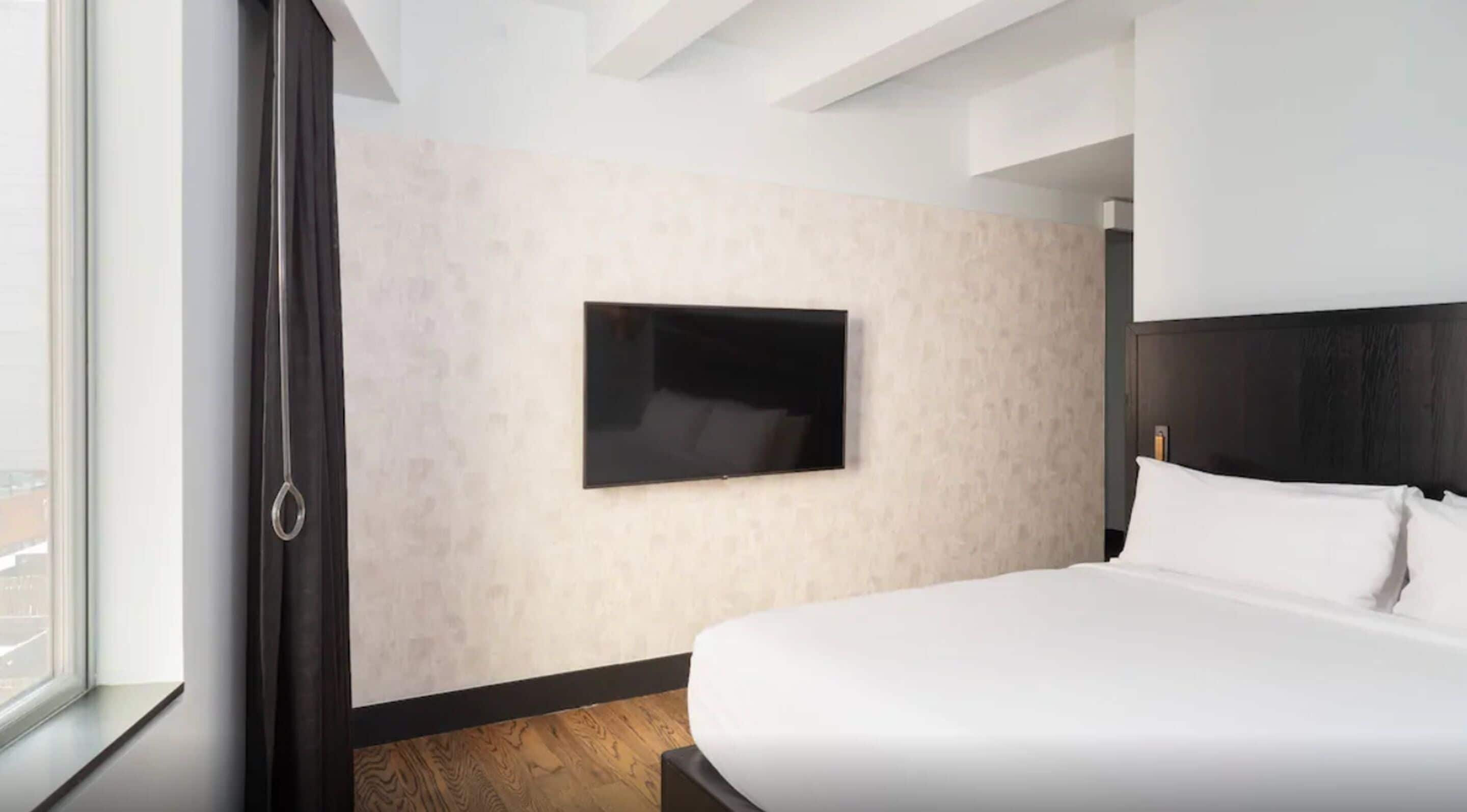 Habitación de hotel moderna con una cama grande, un televisor de pantalla plana montado en la pared y vistas a la ciudad desde la ventana.