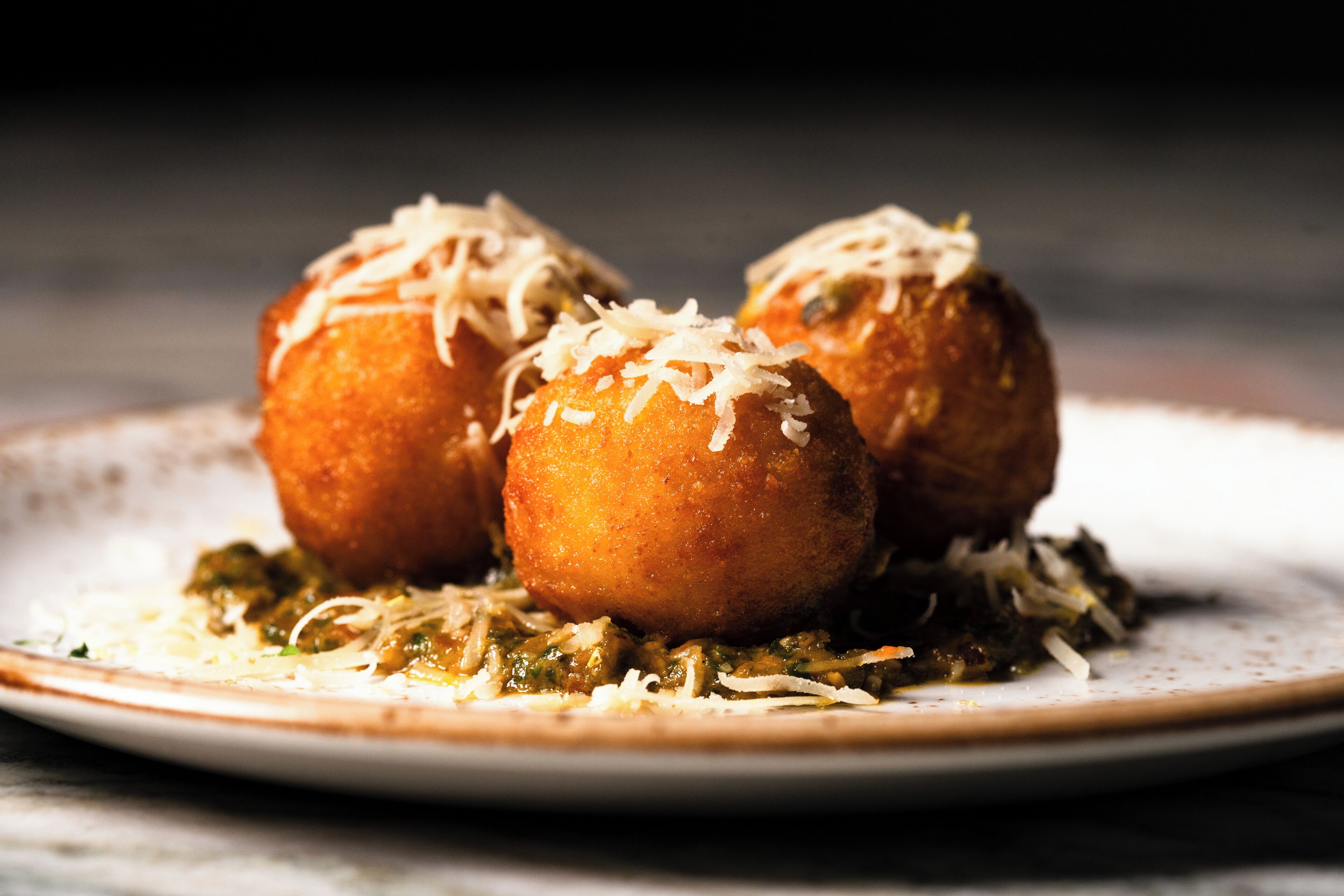 Tres bolas fritas redondas y doradas sobre un plato, con queso rallado encima, acompañadas de una salsa verde.
