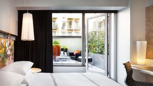 A modern hotel room with a bed, a desk, a chair, a hanging light fixture, and a balcony with outdoor furniture and plants.