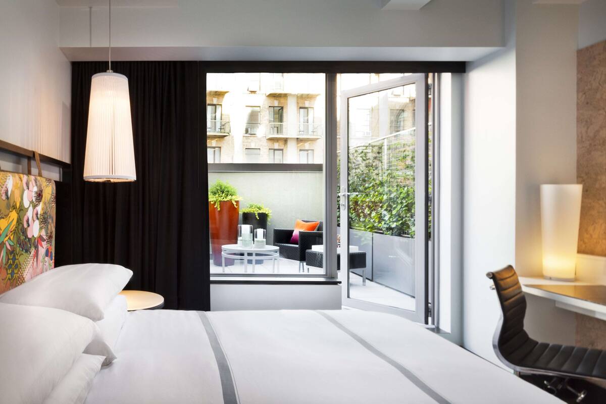 Una habitación de hotel moderna con cama, escritorio, silla, lámpara colgante y balcón con mobiliario exterior y plantas.