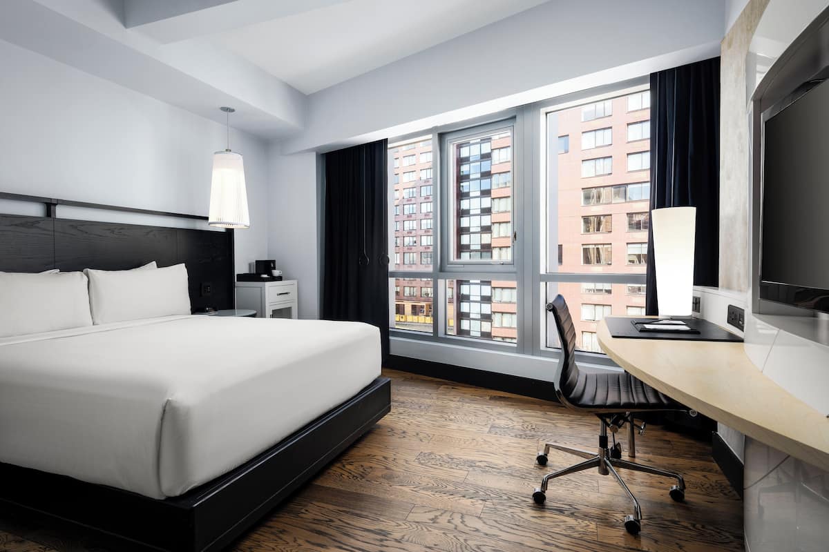 Una habitación de hotel moderna con una cama grande, un escritorio, una silla y un ventanal con vistas a la ciudad.