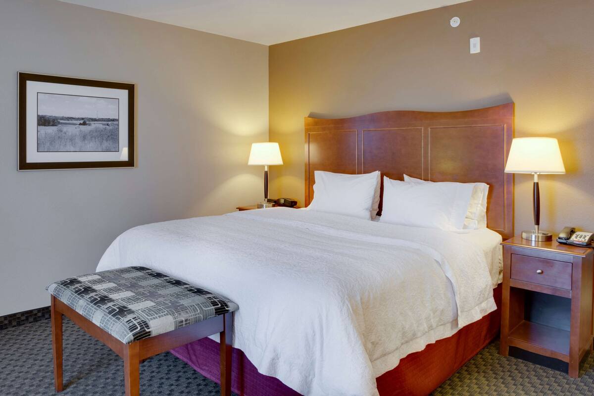 A hotel room with a large bed, two bedside lamps, a bedside table with a lamp, a framed picture on the wall, and a bench at the foot of the bed.