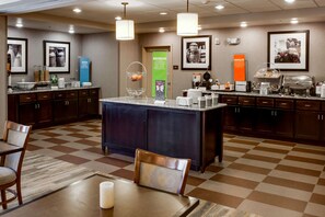 A hotel lobby with a checkered floor, a central island with a coffee station, and a seating area with a table and chairs.