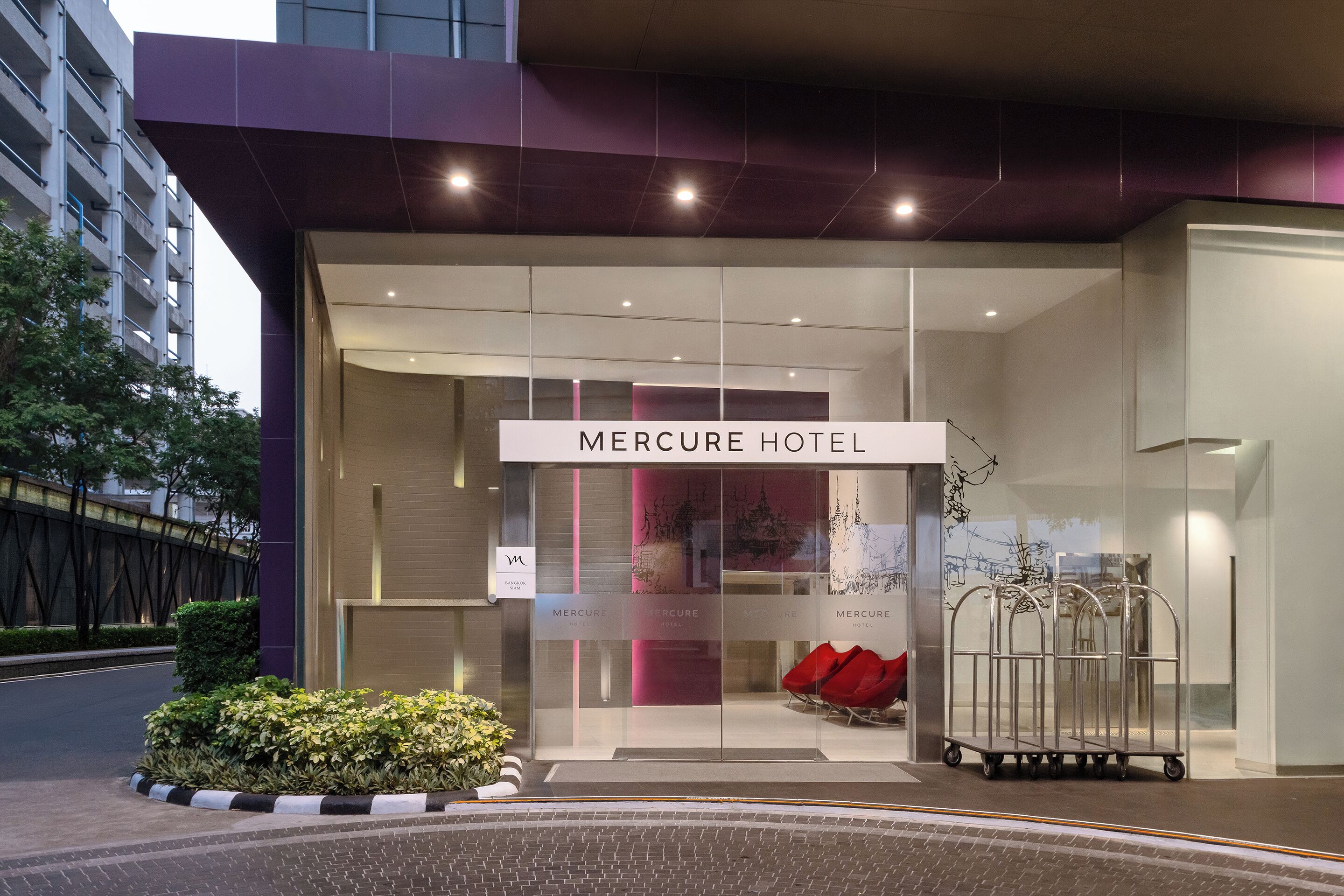 El exterior del Hotel Mercure, con fachada de vidrio, un cartel y un carrito metálico cerca de la entrada.