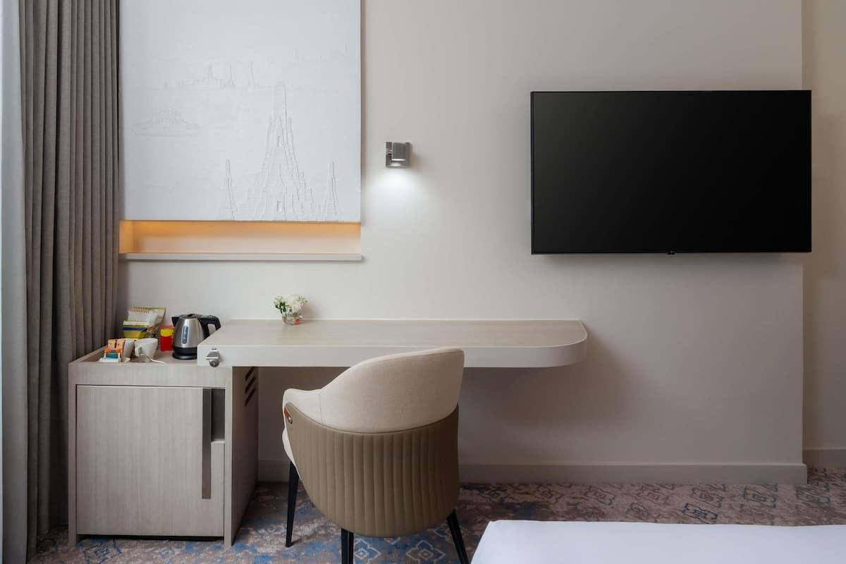 A hotel room with a bed, a desk with a chair, a flat-screen TV, a wall-mounted artwork, and a small table with a vase of flowers.