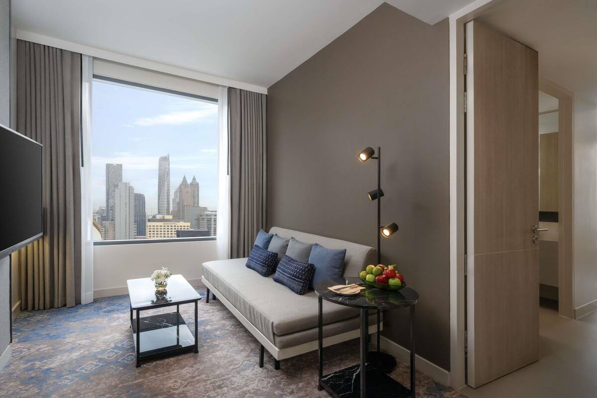 A modern hotel room with a large window, a sofa, a small table, and a floor lamp.