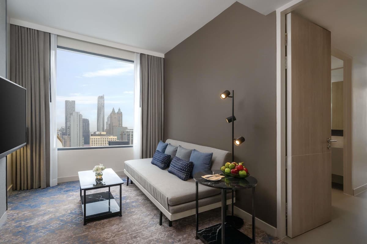 A modern hotel room with a large window, a sofa, a small table, and a floor lamp.